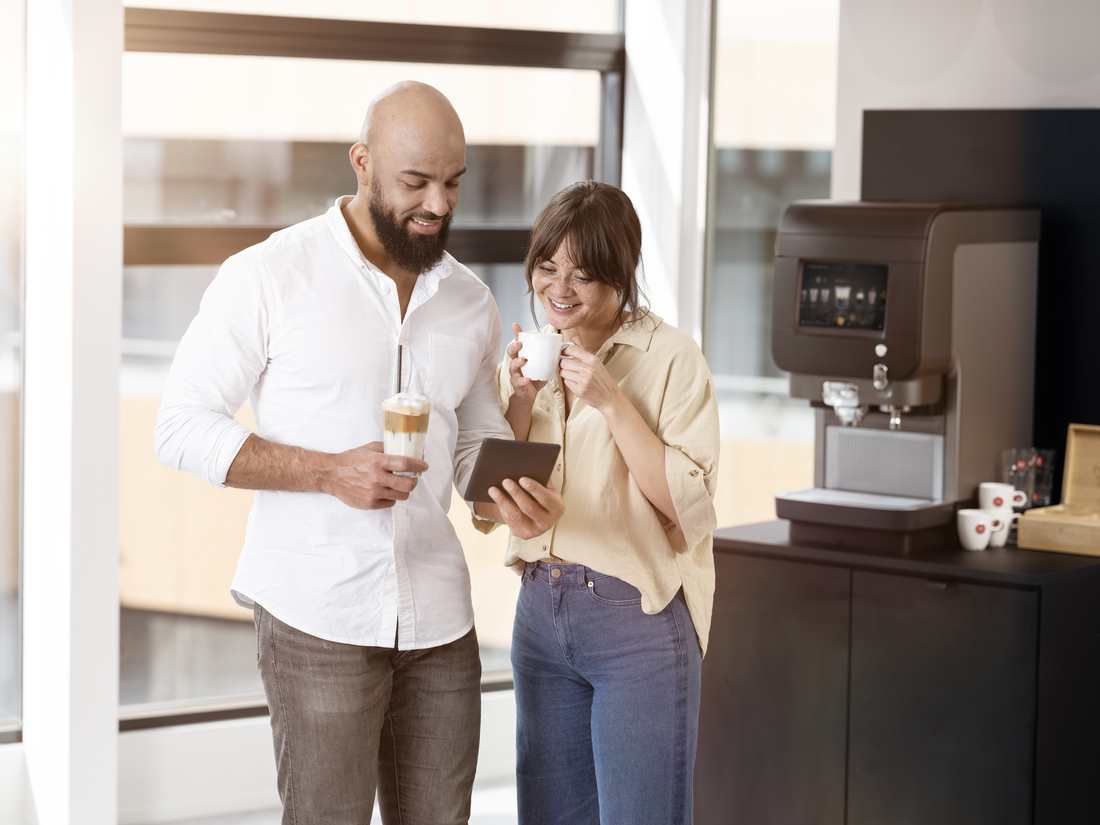 Two people conferring at a coffee machine with a tablet and drinks in a bright office environment.