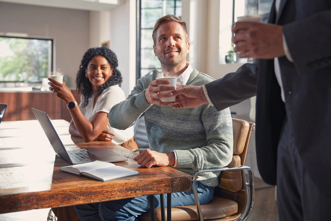 Three colleagues in a bright meeting room, one offering coffee while the others discuss matters with smiles on their faces.